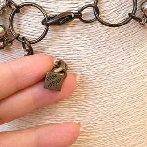 Puede incluir: Primer plano de un collar con un cierre y eslabones de cadena de color bronce. El collar presenta peque&ntilde;as cuentas de color plateado. Un peque&ntilde;o dije cuadrado con el texto "PAM MIRAN" est&aacute; unido a la cadena.