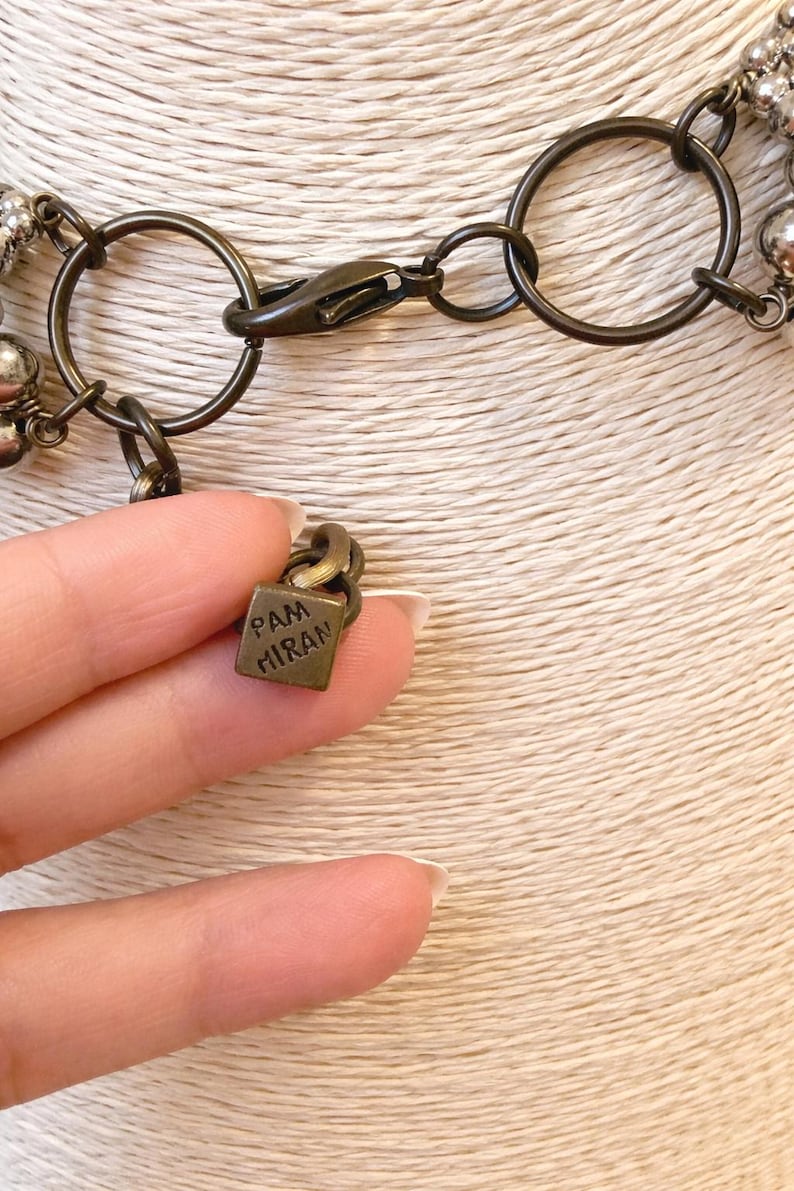 Puede incluir: Primer plano de un collar con un cierre y eslabones de cadena de color bronce. El collar presenta peque&ntilde;as cuentas de color plateado. Un peque&ntilde;o dije cuadrado con el texto "PAM MIRAN" est&aacute; unido a la cadena.