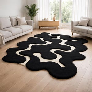 May include: A large, abstract rug with a black, amoeba-like design against a cream-coloured background. The rug is placed on a light-coloured hardwood floor in a living room setting. The rug's dimensions are approximately 244 cm by 152 cm.