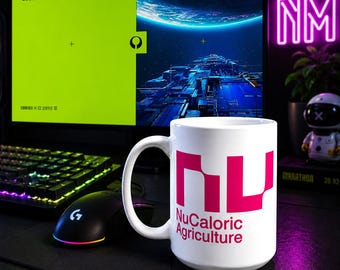 May include: A white ceramic mug featuring a bold pink logo with the stylized letters NU and the text NuCaloric Agriculture printed in pink below. The mug has a curved handle and is positioned in front of a computer monitor and a black gaming mouse.