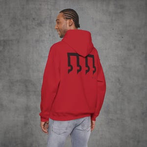 Könnte beinhalten: Ein roter Kapuzenpullover mit einem schwarzen Grafikdesign auf dem Rücken. Das Design ist ein stilisiertes Symbol. Der Kapuzenpullover hat eine Kapuze und lange Ärmel. Die Person im Bild trägt eine blaue Jeans.