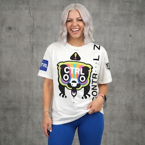 Puede incluir: Camiseta blanca con un gráfico colorido de un personaje de dibujos animados con el texto "CTRL" y "CTRL + L". La camiseta tiene un cuadrado azul con "TRL" en la manga izquierda. La camiseta se combina con leggings azules.