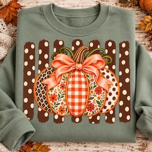 May include: A sage green crewneck sweatshirt featuring a central graphic of a pumpkin adorned with a large peach-colored bow. The pumpkin segments display varied patterns including leopard print, orange gingham, and floral motifs, set against brown vertical stripes.