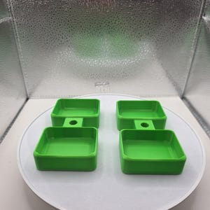 May include: Four bright green, square-shaped trays with a small circular hole on one side are arranged on a white, circular platform. The trays have a simple, modern design and are evenly spaced. The background is a reflective, silver surface.