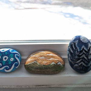 Puede incluir: Tres piedras pintadas con diferentes diseños. Una es azul con remolinos blancos, otra representa una escena de montaña con tonos naranja y verde, y la tercera es azul oscuro con un patrón en zigzag blanco.
