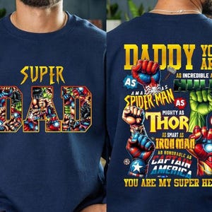 May include: Navy blue sweatshirt featuring the text "SUPER DAD" in gold, filled with comic book characters. The back of the sweatshirt says "DADDY YOU ARE MY SUPER HERO" with superhero references.