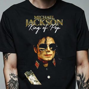 May include: Black t-shirt with a portrait of Michael Jackson wearing sunglasses. The text "MICHAEL JACKSON King of Pop" is printed in gold above the image. The shirt has a classic crew neck design.