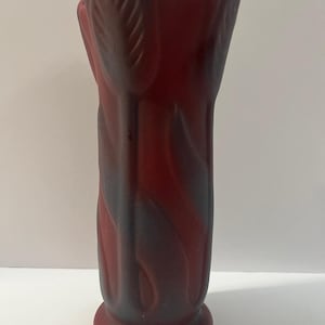 May include: A tall, cylindrical vase with a matte, deep red finish. The exterior features a raised, molded relief pattern of vertical tulip flowers and stems that extend from the base to the scalloped, petal-shaped rim at the top of the vessel.