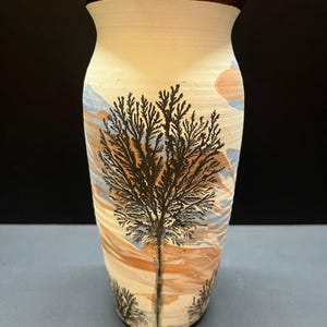 May include: A ceramic vase with a flared rim and a dark brown glazed top edge. The body features a marbled pattern of cream, tan, and light blue swirls, overlaid with a black, branching, tree-like dendrite design on the front surface.