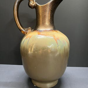 May include: A ceramic pitcher featuring a glossy finish with a gradient of muted olive green and brown tones. The upper neck and handle are finished in a metallic bronze glaze, while the body displays subtle orange and blue drip patterns.