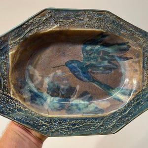 May include: An octagonal ceramic bowl featuring a hand-painted blue hummingbird in flight against a brown and tan background. The wide, textured rim is finished in a deep blue glaze with a crackled, weathered appearance and dark edges.