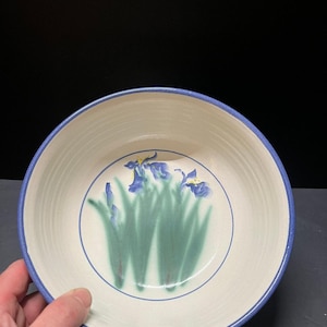 May include: A round ceramic bowl with a cream-colored interior and a blue rim. The center of the bowl features a hand-painted design of blue iris flowers with green stems and leaves, enclosed within a thin blue circular line on the base.