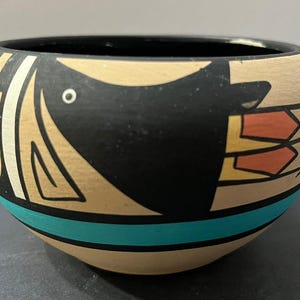 May include: A rounded ceramic bowl featuring a tan base color with a black, turquoise, and orange graphic design. The exterior displays a stylized black bird figure with an orange and yellow arrow-like tail, set against a horizontal turquoise band.