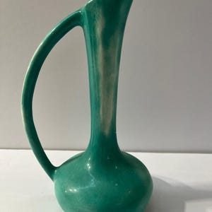 May include: A tall, slender ceramic pitcher with a vibrant teal green glaze and white highlights. The vessel features a rounded base, a long, narrow neck that flares slightly at the top, and a single curved handle extending from the neck.