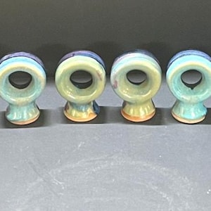 May include: A set of twelve circular ceramic napkin rings arranged in a semi-circle. Each ring features a glossy glaze with a blend of light blue, pale green, and hints of purple, with a visible terracotta-colored base on the bottom of each piece.