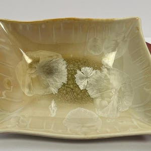 May include: A rectangular ceramic dish with a wavy rim, featuring a glossy cream-colored glaze. The center displays a textured, crackled olive-green pattern surrounded by several white, starburst-like crystalline formations.