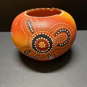 May include: A round, hollowed-out gourd bowl featuring a vibrant orange and yellow gradient base. The exterior is decorated with intricate dot-painted patterns in black, white, and gold, forming circular and crescent shapes around the surface.