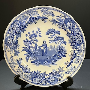May include: A round ceramic plate featuring a blue and white transferware pattern. The center depicts a person standing by a well in a landscape, while the border is decorated with intricate floral motifs and foliage on a cream-colored background.