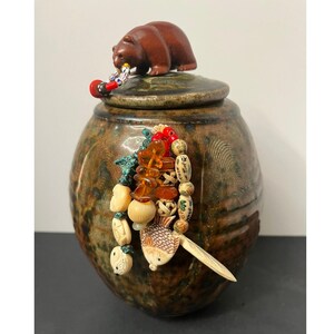 May include: A round, mottled brown ceramic jar with a lid topped by a small brown bear figurine. A decorative cluster of beads, including amber-colored pieces, carved bone-like fish shapes, and small red accents, hangs from the side of the jar.
