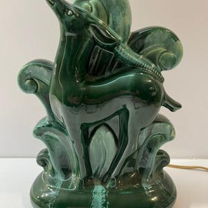 May include: A glossy dark green ceramic lamp shaped like a standing deer. The deer is positioned in front of stylized, curved foliage elements. The glaze features variations in tone, ranging from deep forest green to lighter teal highlights.
