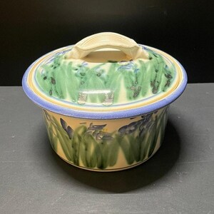 May include: A round ceramic lidded jar featuring a hand-painted design of green leaves and blue iris flowers on a cream-colored background. The lid has a sculpted handle and a blue rim, while the base also features a matching blue painted rim.
