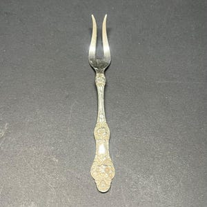 May include: A silver-toned metal cocktail fork featuring two long, slender tines and a decorative handle with an intricate embossed floral pattern. The utensil has a polished finish and a tapered neck leading to a wider, ornate base with detailed relief.