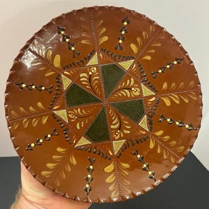 May include: A round, brown ceramic plate featuring a scalloped edge and a central geometric star pattern in dark green and light yellow. The surface is decorated with hand-painted floral motifs in yellow and black, arranged in a radial design.
