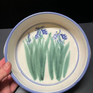 May include: A round ceramic bowl with a cream-colored interior featuring a hand-painted design of green iris leaves and blue iris flowers. The bowl has a blue rim and a thin blue circular line accenting the interior base of the vessel.