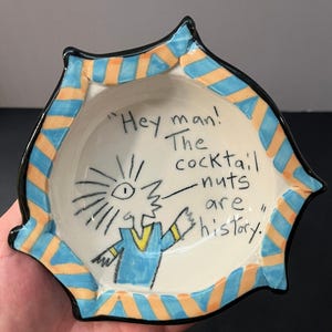 May include: A ceramic bowl with a wavy, scalloped rim featuring blue and orange diagonal stripes. The interior is off-white with a hand-drawn illustration of a character and the text: "Hey man! The cocktail nuts are history."