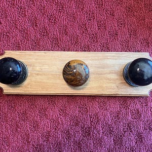 May include: A wooden coat rack with three decorative knobs. The knobs are black and brown, and the wood has a light finish. The rack is rectangular with rounded edges. The knobs are evenly spaced along the length of the rack.