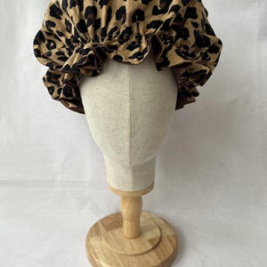 Op de afbeelding: Een haarkap met een beige en zwart luipaardpatroon. De kap heeft een geplooide, gegolfde rand en een brede, elastische band die ontworpen is om stevig rond het hoofd te zitten, met een gladde textuur over het gehele ontwerp.