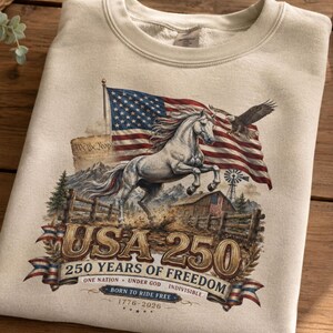 May include: A beige crewneck sweatshirt featuring a graphic of a white horse, an American flag, a bald eagle, a barn, and mountains. The text reads USA 250, 250 YEARS OF FREEDOM, ONE NATION UNDER GOD INDIVISIBLE, BORN TO RIDE FREE, and 1776-2026.