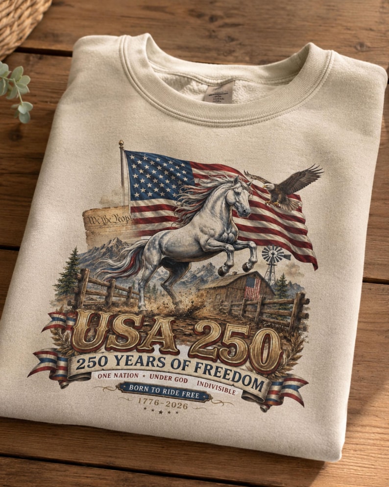 May include: A beige crewneck sweatshirt featuring a graphic of a white horse, an American flag, a bald eagle, a barn, and mountains. The text reads USA 250, 250 YEARS OF FREEDOM, ONE NATION UNDER GOD INDIVISIBLE, BORN TO RIDE FREE, and 1776-2026.