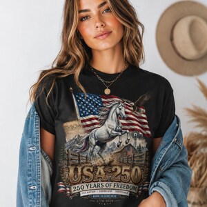 May include: A black t-shirt featuring a graphic of a rearing white horse, an American flag, a bald eagle, a barn, and a windmill. The text reads USA 250, 250 YEARS OF FREEDOM, ONE NATION UNDER GOD INDIVISIBLE, BORN TO RIDE FREE, and 1776-2026.