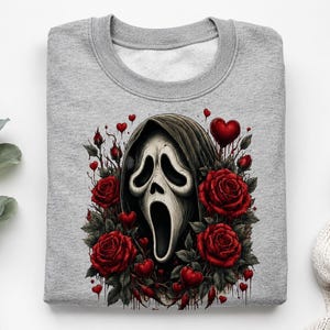 May include: A heather gray crewneck sweatshirt featuring a central graphic of a white ghost mask with a dark hood, surrounded by deep red roses and small red hearts. The design includes dark, dripping accents around the floral elements.