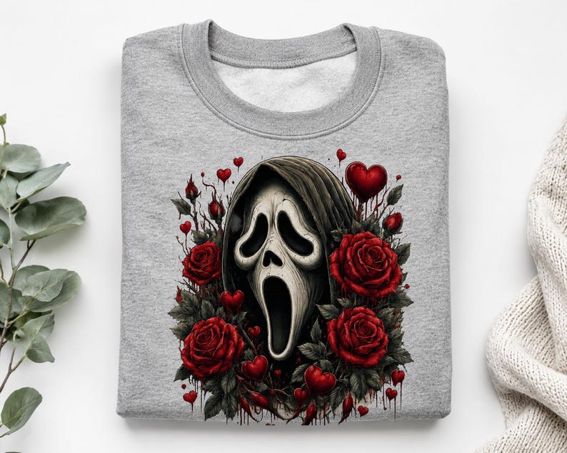 May include: A heather gray crewneck sweatshirt featuring a central graphic of a white ghost mask with a dark hood, surrounded by deep red roses and small red hearts. The design includes dark, dripping accents around the floral elements.
