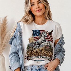May include: A white t-shirt featuring a graphic print of a rearing white horse, an American flag, a bald eagle, a barn, and the text USA 250, 250 YEARS OF FREEDOM, ONE NATION UNDER GOD INDIVISIBLE, BORN TO RIDE FREE, and the years 1776-2026.