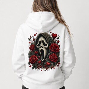 May include: A white hooded sweatshirt featuring a large graphic on the back. The design displays a central ghost-like mask with a dark hood, surrounded by deep red roses, green leaves, and small red hearts, with subtle dripping paint effects.