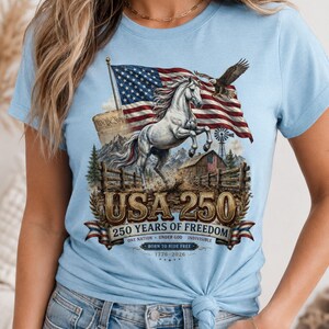 May include: A light blue t-shirt featuring a graphic print of a rearing white horse, an American flag, a bald eagle, a barn, and mountains. The text reads USA 250, 250 YEARS OF FREEDOM, ONE NATION UNDER GOD INDIVISIBLE, BORN TO RIDE FREE, 1776-2026.