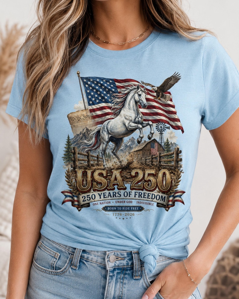 May include: A light blue t-shirt featuring a graphic print of a rearing white horse, an American flag, a bald eagle, a barn, and mountains. The text reads USA 250, 250 YEARS OF FREEDOM, ONE NATION UNDER GOD INDIVISIBLE, BORN TO RIDE FREE, 1776-2026.