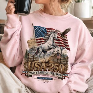 May include: A light pink crewneck sweatshirt featuring a graphic print of a white horse, an American flag, a bald eagle, a barn, and the text USA 250 250 YEARS OF FREEDOM ONE NATION UNDER GOD INDIVISIBLE BORN TO RIDE FREE 1776-2026.