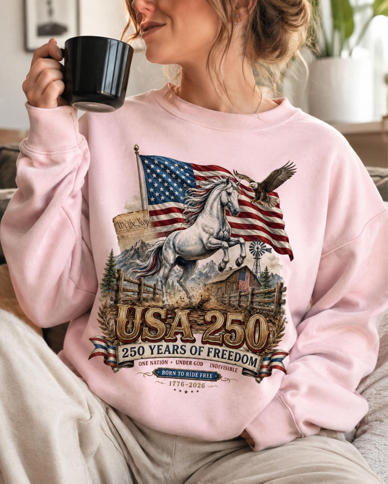 May include: A light pink crewneck sweatshirt featuring a graphic print of a white horse, an American flag, a bald eagle, a barn, and the text USA 250 250 YEARS OF FREEDOM ONE NATION UNDER GOD INDIVISIBLE BORN TO RIDE FREE 1776-2026.