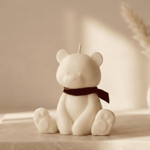以下が含まれることがあります： 濃い茶色のスカーフをつけた、白いクマの形をしたキャンドル。キャンドルは上部に芯があり、明るい色の表面に置かれています。クマは足を広げて座っています。背景には植物が入った装飾的な花瓶があります。