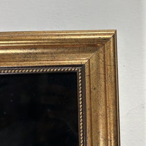 Op de afbeelding: Een rechthoekige fotolijst met een verweerde gouden afwerking en een donkerbruine binnenrand. De binnenrand is versierd met een rij kleine, verhoogde gouden kralen. De lijst omsluit een effen zwart middenpaneel, wat zorgt voor een opvallend contrast.