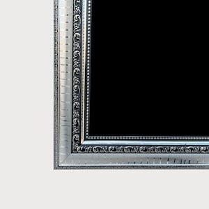 Op de afbeelding: Zilveren fotolijst met sierlijke details. De lijst heeft een rechthoekige vorm met een zwart interieur. De rand van de lijst heeft een herhalend patroon en een parelrand, wat een klassieke en elegante uitstraling creëert.