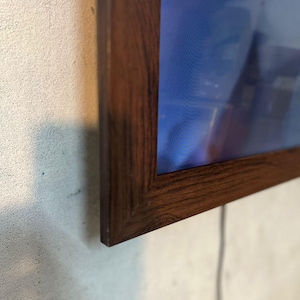 Può includere: Una cornice rettangolare in legno marrone scuro con un motivo a venature del legno visibile. La cornice circonda uno schermo blu e un cavo bianco si estende dal basso. La cornice è montata su un muro grigio chiaro strutturato.