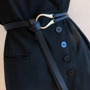 Puede incluir: Un cintur&oacute;n negro con una hebilla dorada &uacute;nica envuelve una blazer negra. El dise&ntilde;o del cintur&oacute;n presenta un lazo y una correa delgada que cuelga. La blazer tiene botones negros y un bolsillo.