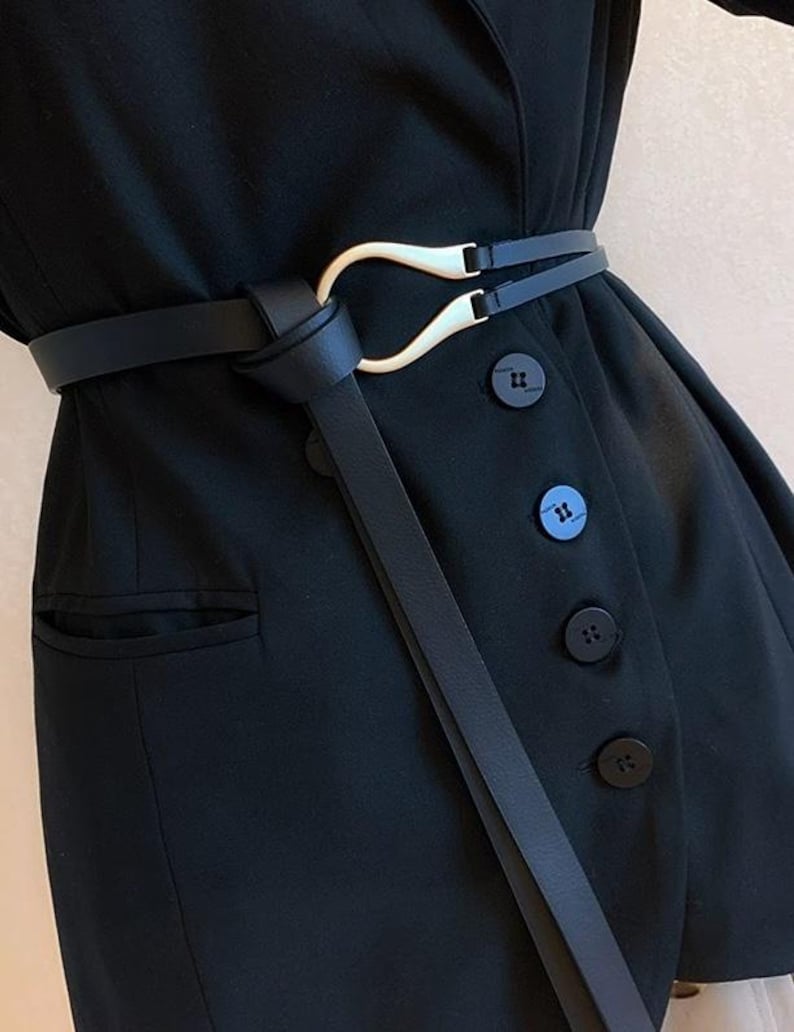 Puede incluir: Un cintur&oacute;n negro con una hebilla dorada &uacute;nica envuelve una blazer negra. El dise&ntilde;o del cintur&oacute;n presenta un lazo y una correa delgada que cuelga. La blazer tiene botones negros y un bolsillo.