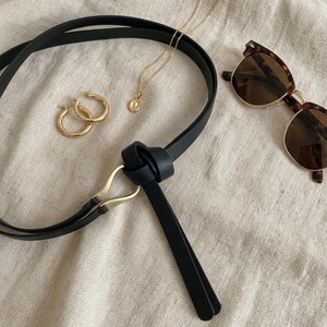 Puede incluir: Un surtido de accesorios que incluye un cintur&oacute;n de cuero negro con hebilla dorada, pendientes de aro dorados, un collar con colgante dorado y gafas de sol carey. Los art&iacute;culos est&aacute;n colocados sobre una tela de color neutro.