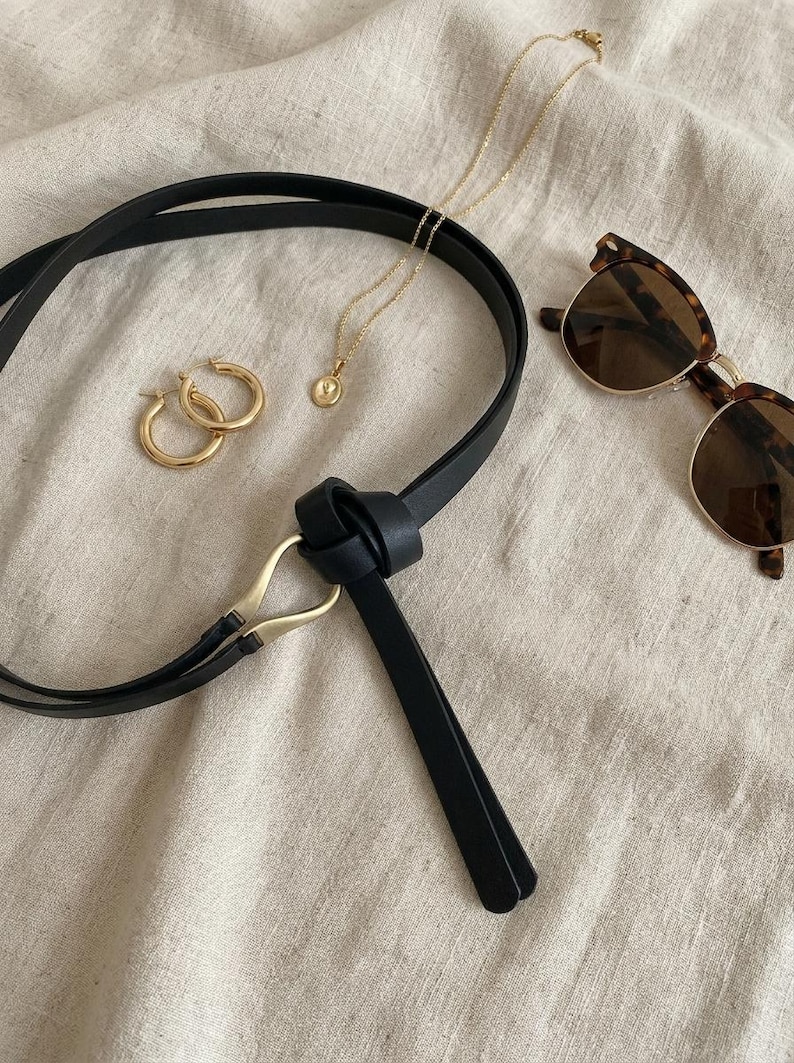 Puede incluir: Un surtido de accesorios que incluye un cintur&oacute;n de cuero negro con hebilla dorada, pendientes de aro dorados, un collar con colgante dorado y gafas de sol carey. Los art&iacute;culos est&aacute;n colocados sobre una tela de color neutro.
